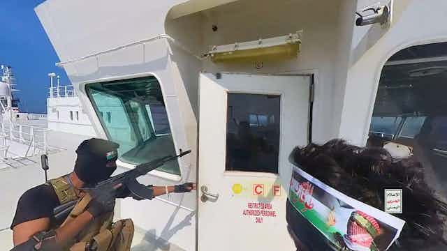 An armed man in military uniform carrying an assault rifle opens the door to the bridge of a cargo vessel.