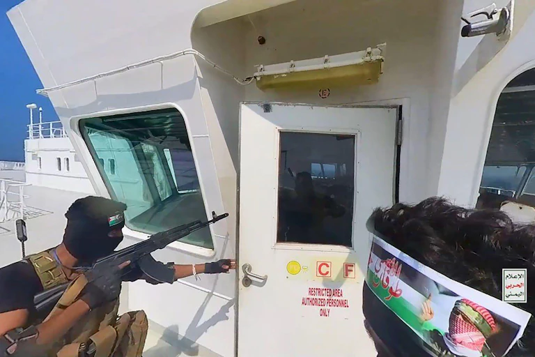 An armed man in military uniform carrying an assault rifle opens the door to the bridge of a cargo vessel.