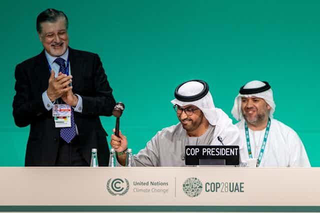 Sultan al-Jaber smiling and about to bring down a gavel. Man in suit stands next to him clapping and smiling.