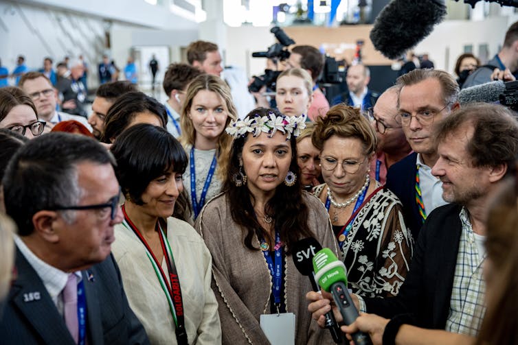 Woman wearing flower garland on her head talks into a microphone, surrounded by other smartly dressed people.