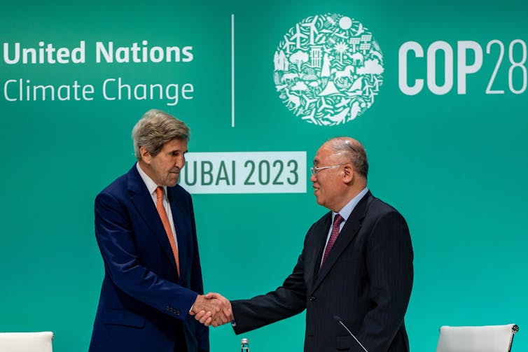 John Kerry and Xie Zhenhua shake hands in front of a sign for COP28.