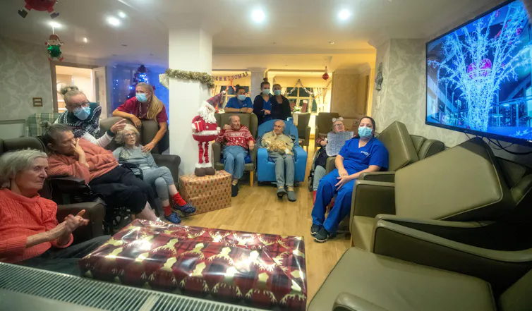 Elderly people and nurses sit in a living room watching TV.