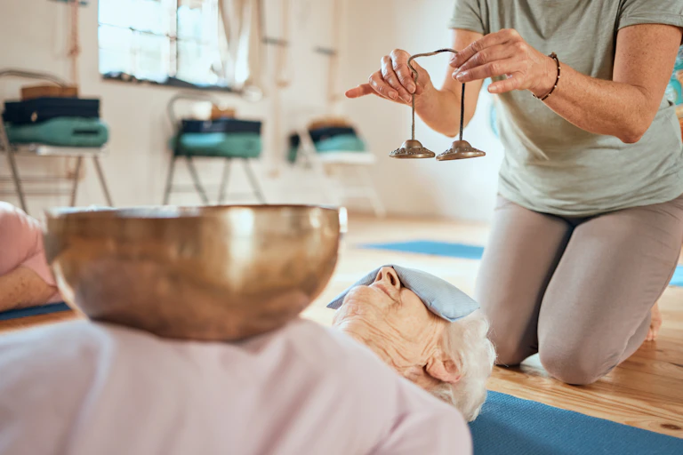 A Tibetan singing bowl rests on the body of a person who is lying down, while another holds bells close to her.