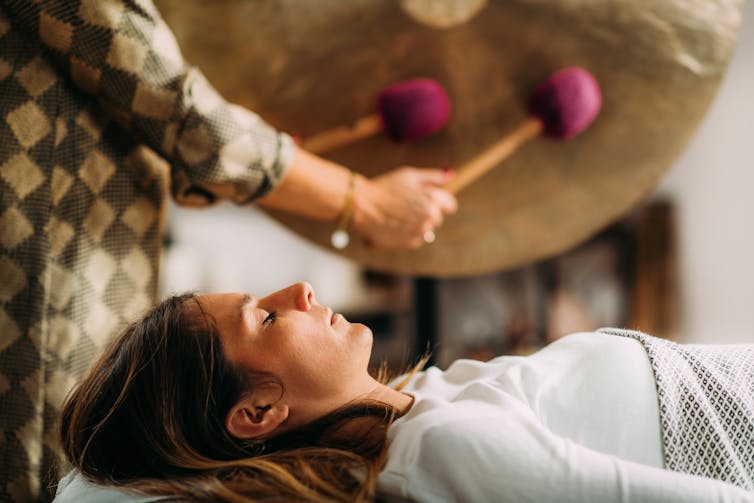 A woman lying down with eyes closed while gongs are played next to her.