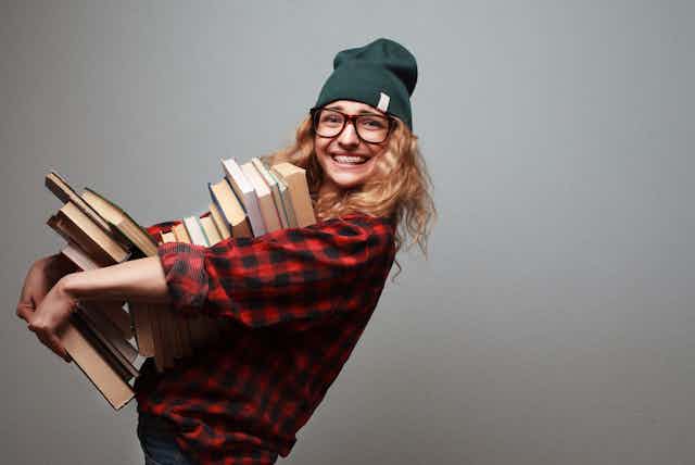 Una adolescente con gafas, gorro de lana y aparato, sonríe mientras sujeta un montón de libros.