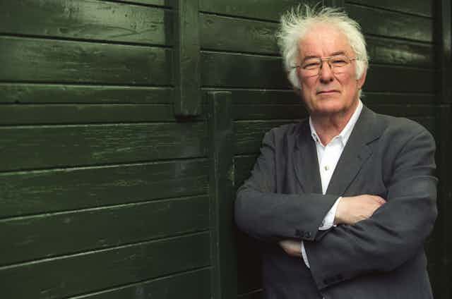 An older man with white hair (Seumas Heaney), arms folded leaning against a wooden fence.