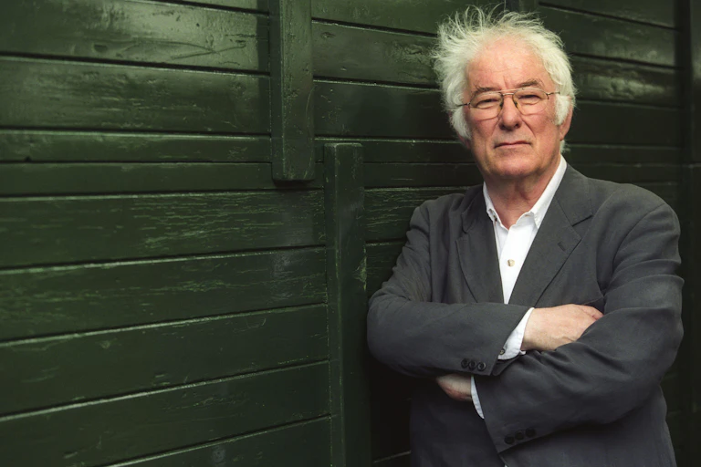 An older man with white hair (Seumas Heaney), arms folded leaning against a wooden fence.