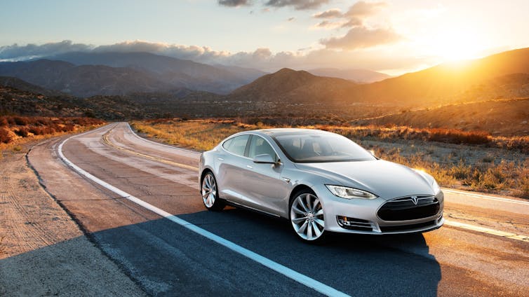 A grey Tesla model S driving on the road with the sun setting in the background.