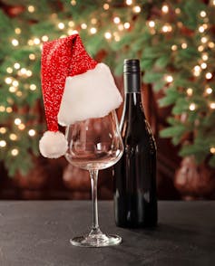 A wine bottle and wine glass wearing a santa hat, with holiday wreath in the background