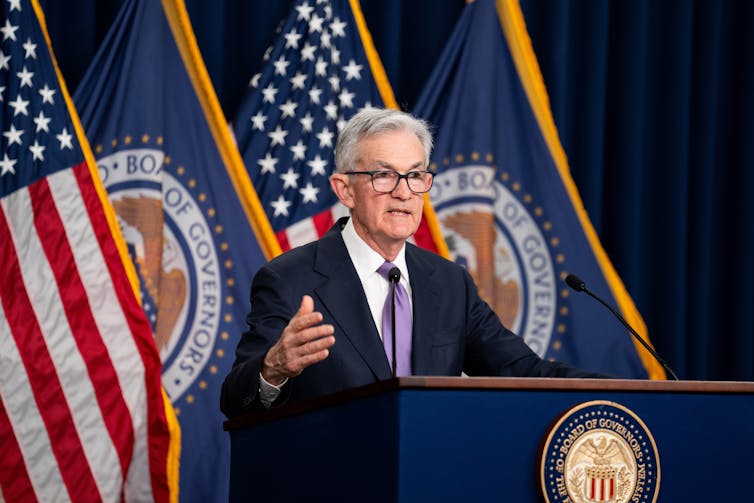 Jay Powell, wearing a suit, gestures toward an unseen crowd during a press conference. He stands behind a podium and in front of a line of flags.