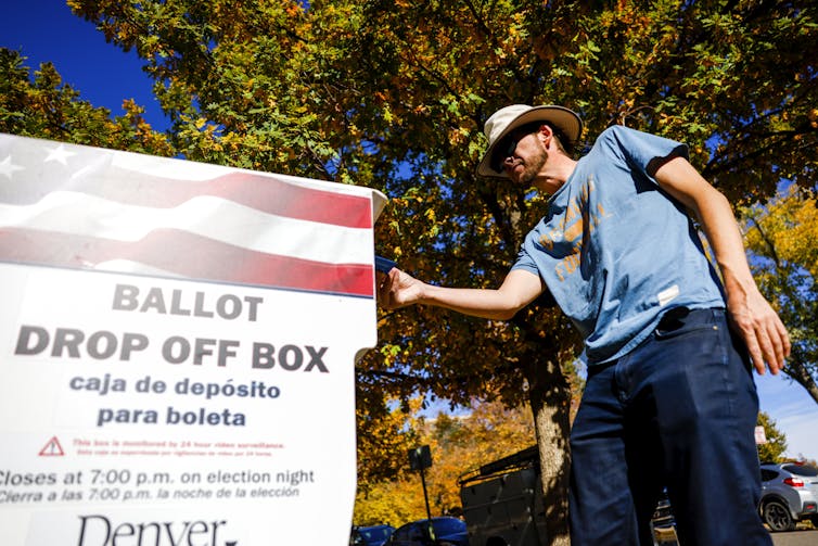 A man wearing a blue shirt and a hat drops off a ballot in an official box.