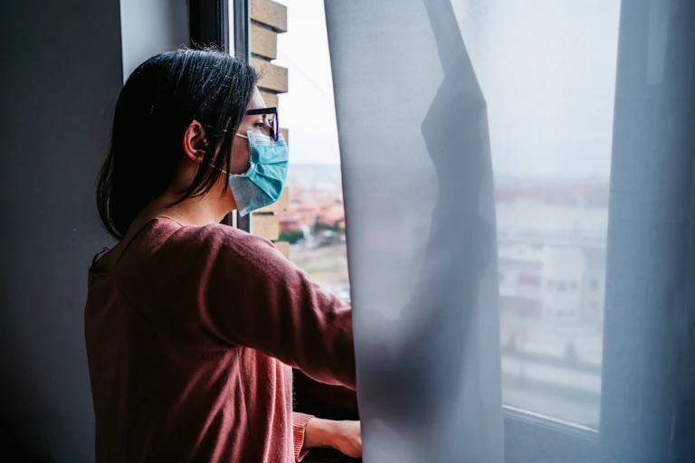 woman in mask looking out a window