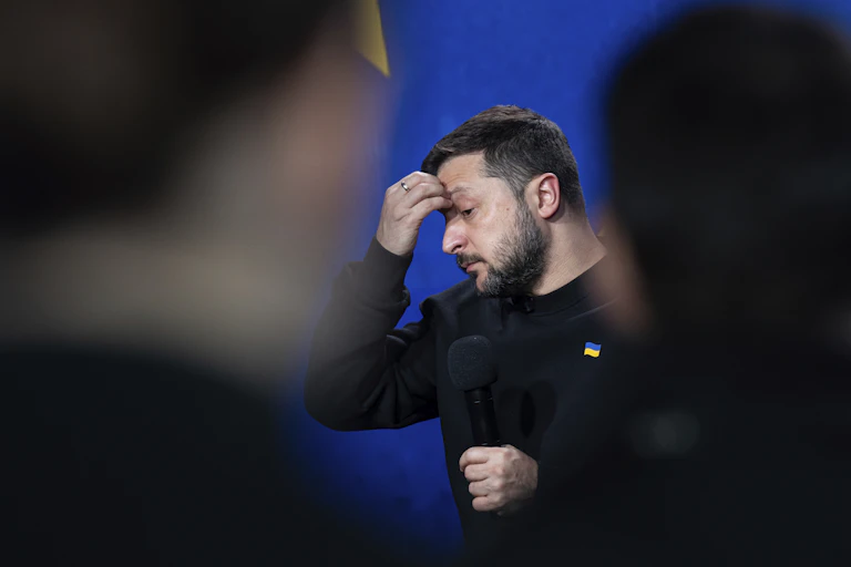 A bearded man with his hand on his forehead photographed between the silhouettes of two reporters.