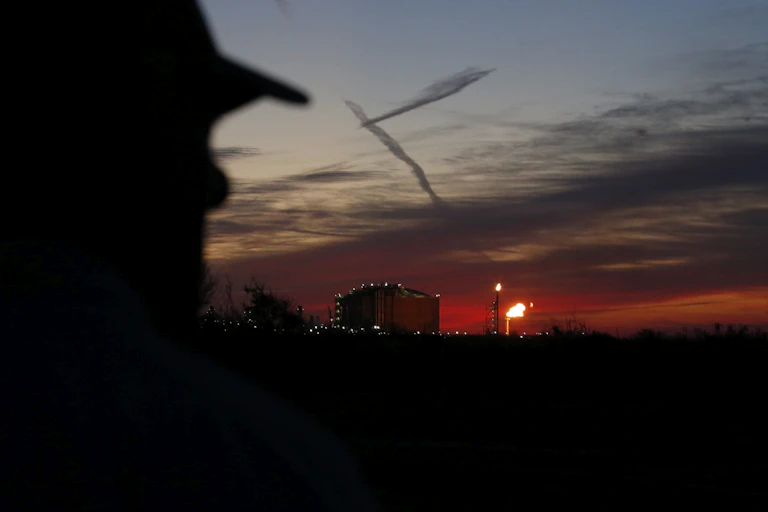 A partially obscured man with a hat is seen looking at a gas flame against a darkening sky.