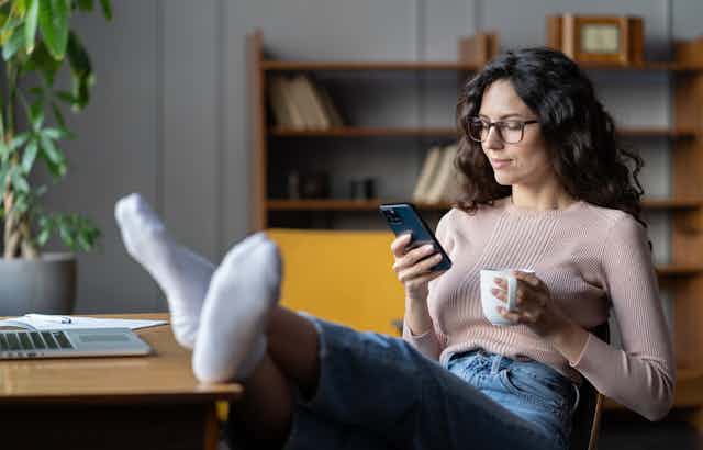 A young woman with dark hair and glasses reads on her mobile phone and holds a cup of coffee, she has her feet up on her desk by a laptop and is in a stylish, modern home office