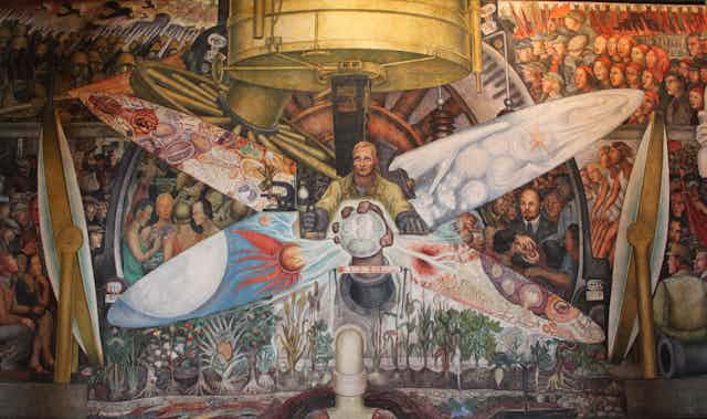 Inmenso mural que retrata a un hombre sosteniendo un engranaje rodeado de gente representando el capitalismo a un lado y el socialismo al otro.