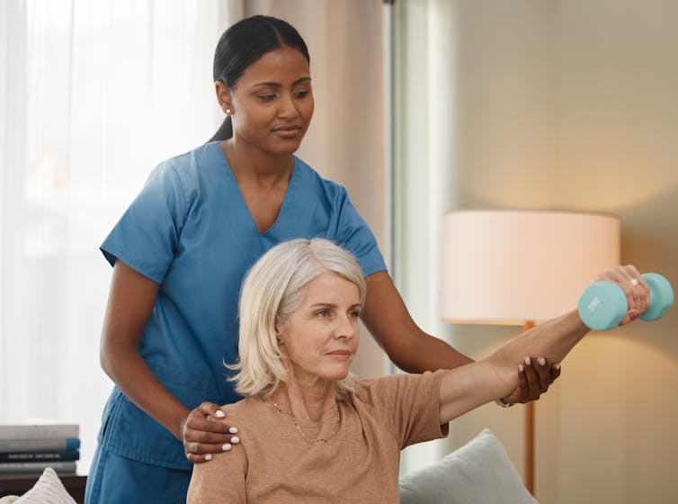 A seated woman lifting a small dumbbell with assistance from a woman in scrubs standing behind her
