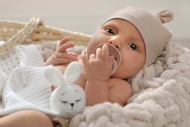 A blue-eyed infant wearing a beige cap and holding a pacifier looks at the camera.