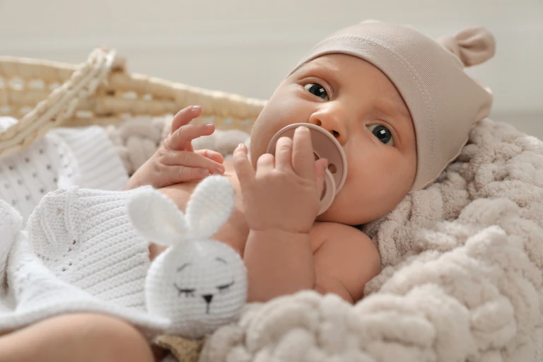 A blue-eyed infant wearing a beige cap and holding a pacifier looks at the camera.