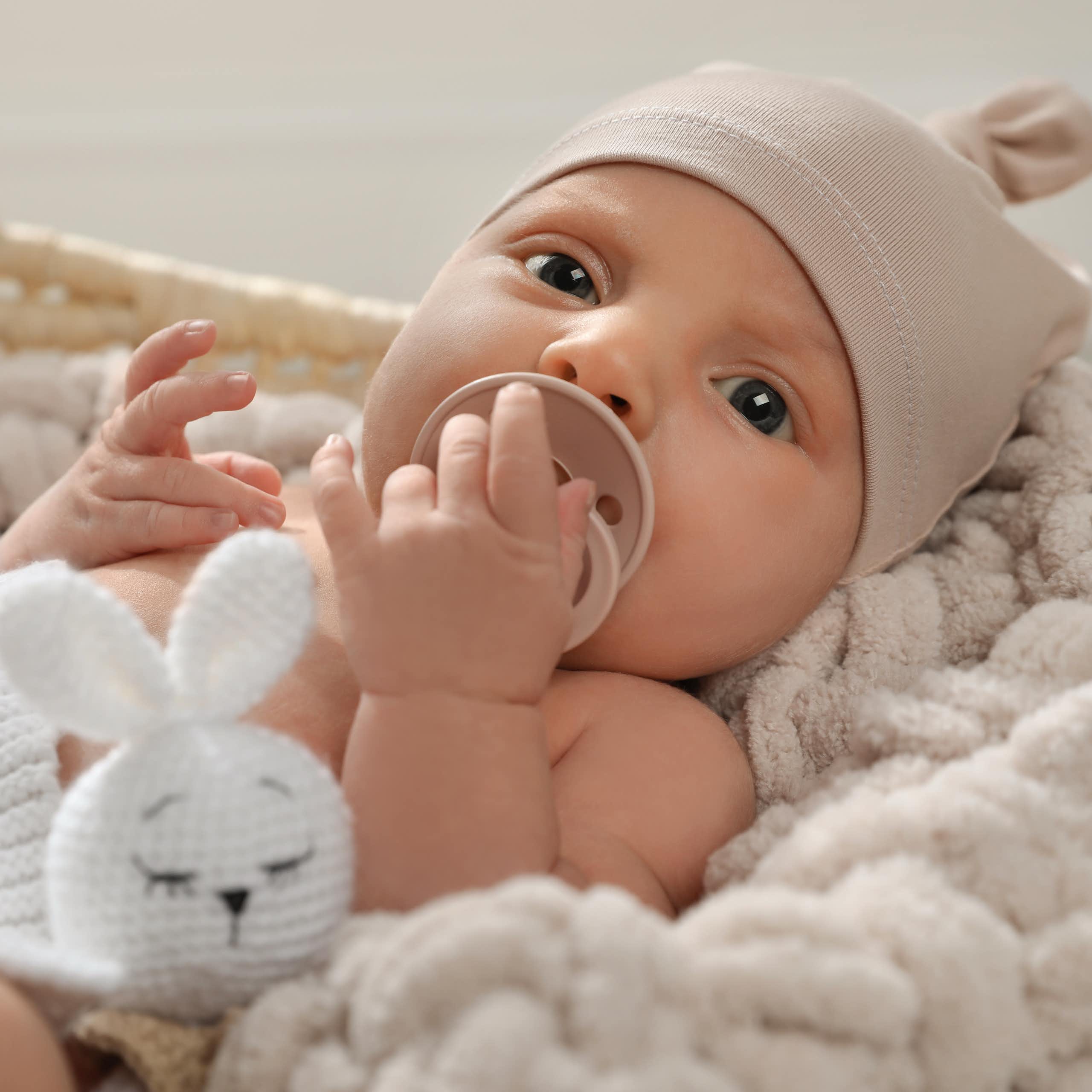 A blue-eyed infant wearing a beige cap and holding a pacifier looks at the camera.