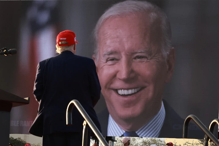 Donald Trump, de dos, pendant un meeting, regarde un écran géant sur lequel on voit Joe Biden.