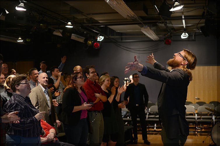 A man with a blonde beard raises his arms and looks upward as he stands facing a small crowd of people in a dimly lit room.