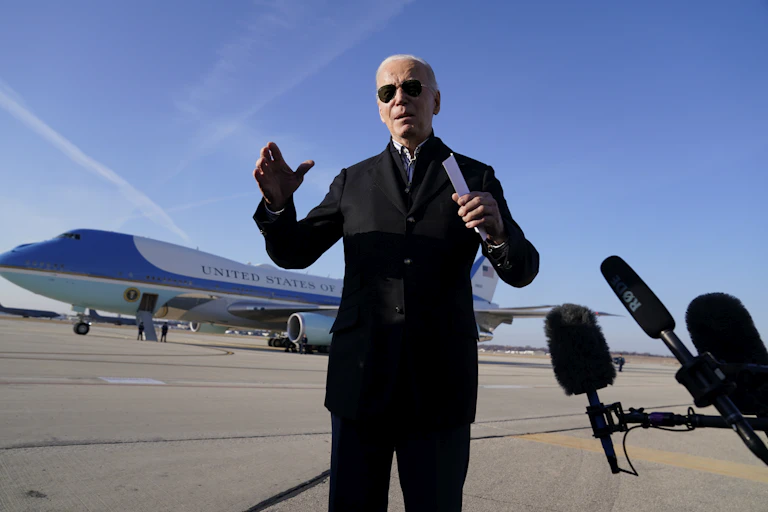 An older man in aviator sunglasses speaks to the media as microphones surround him and a large aircraft sits on a tarmac behind him.