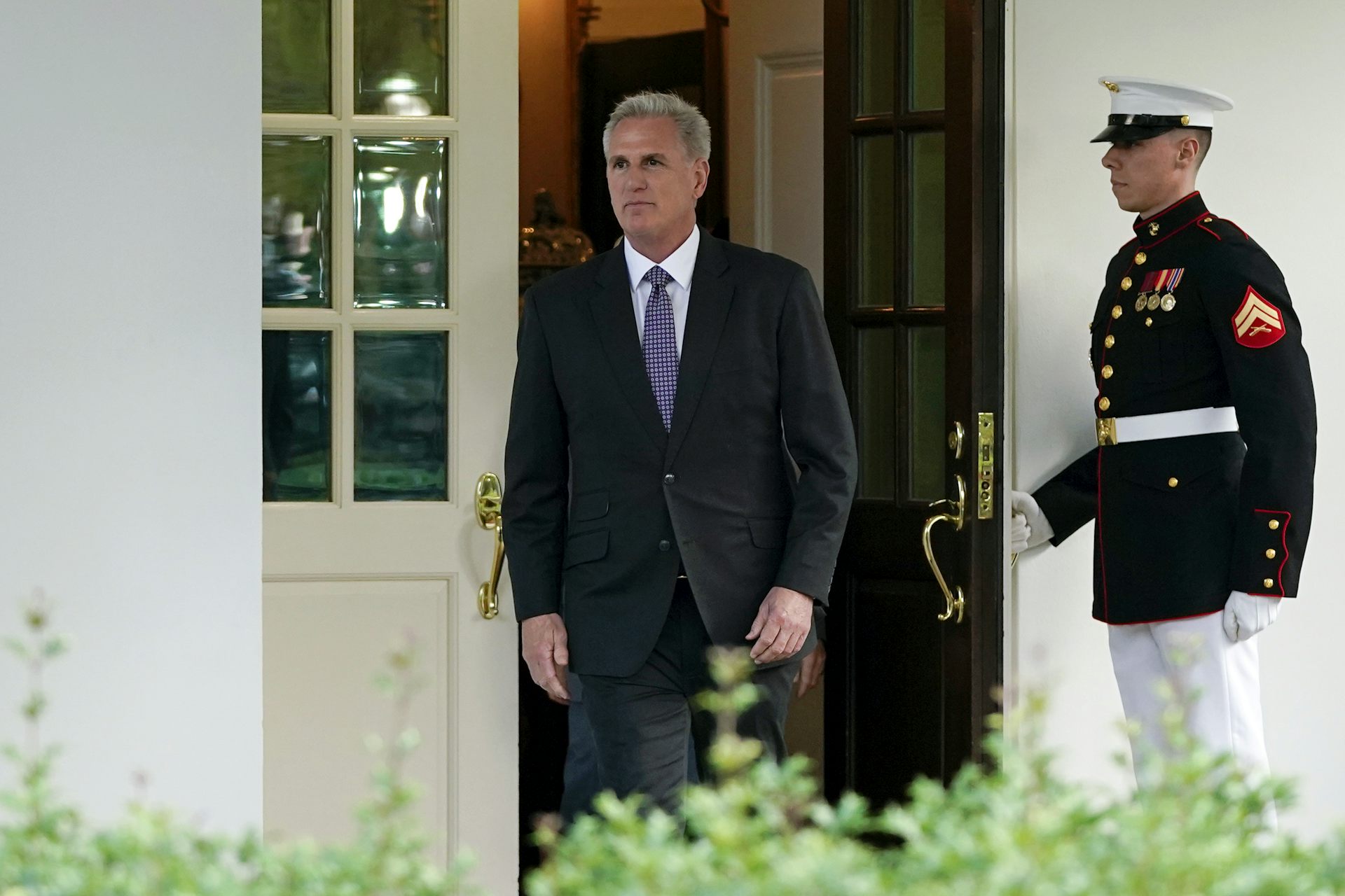 A man in a dark suit walks out of a door as a man in uniform holds it open for him.
