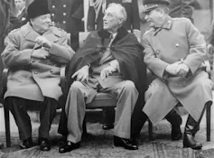A black-and-white photo shows three men in overcoats talking.