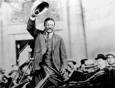 A black and white photo shows a smiling man standing in an automobile with his hat in his raised hand.