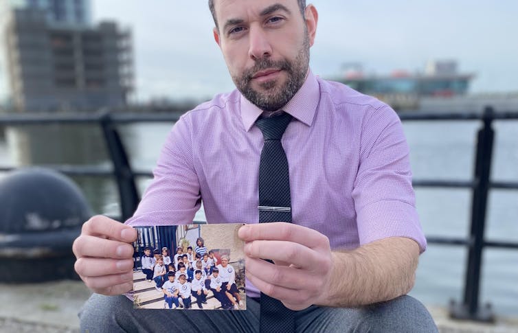 A man in a lilac shirt and tie holds out a photograph.