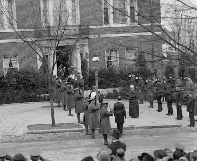 People carry a casket through a front door of a brick building as military officers salute.