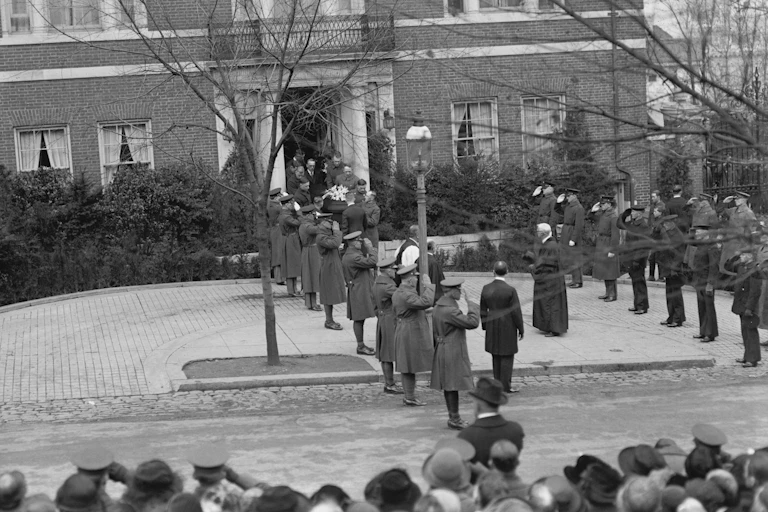 People carry a casket through a front door of a brick building as military officers salute.