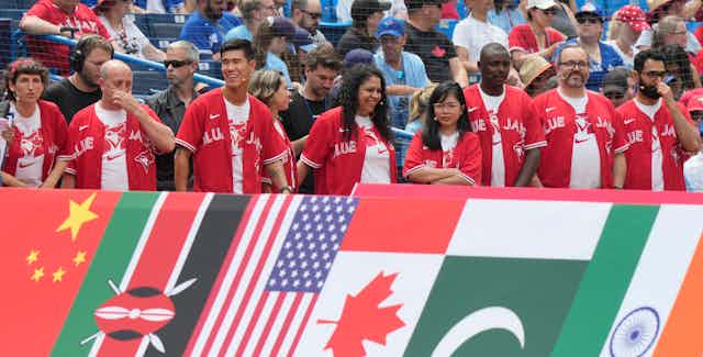 Smiling people in red Blue Jays jersey stand behind a row of flags.