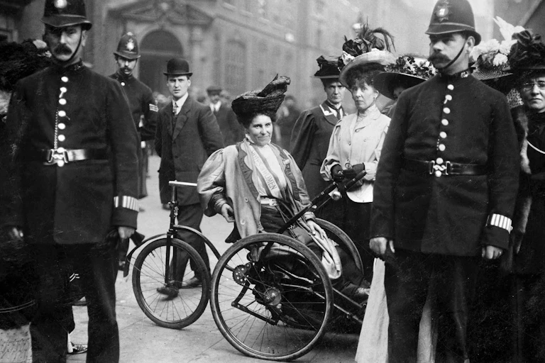 Black and white photo of suffragettes surrounded by police, including Rosa May Billinghurst in her tricycle wheelchair