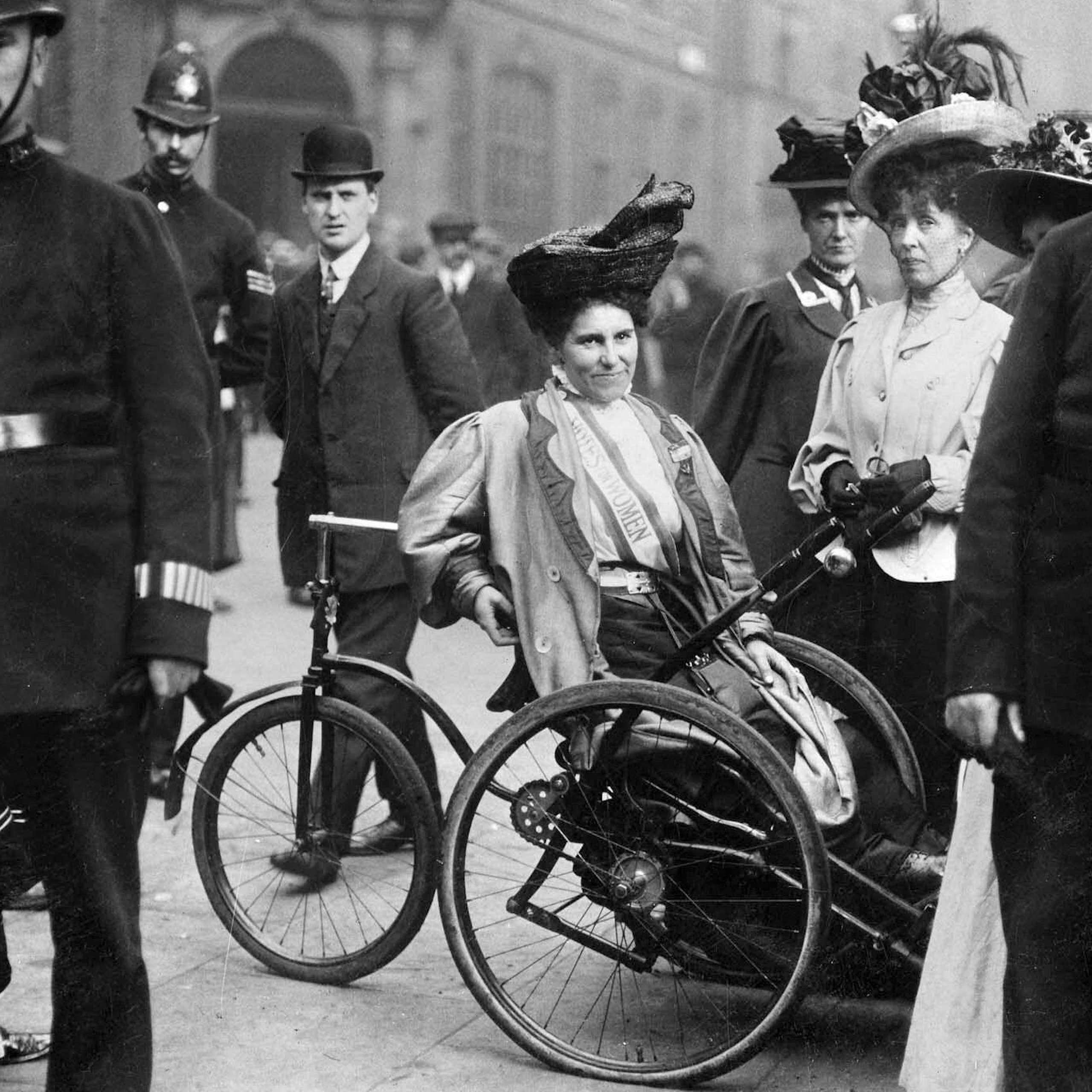 Black and white photo of suffragettes surrounded by police, including Rosa May Billinghurst in her tricycle wheelchair