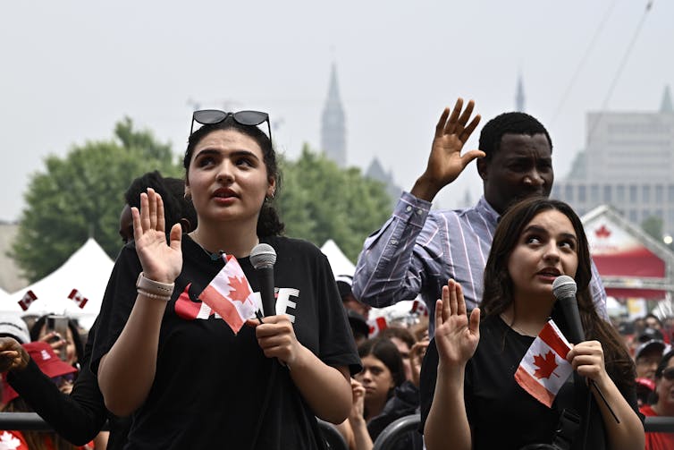 Two women and a man raise their hands and read an oath while holding small Canadian flags.