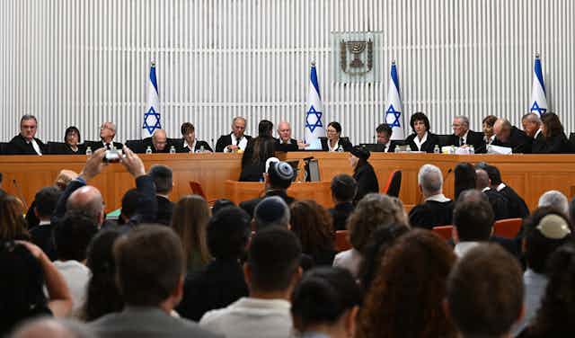 Judges seated at a long desk, backed by blue and white flags, in front of a crowd of seated people.