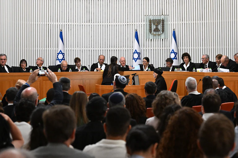 Judges seated at a long desk, backed by blue and white flags, in front of a crowd of seated people.