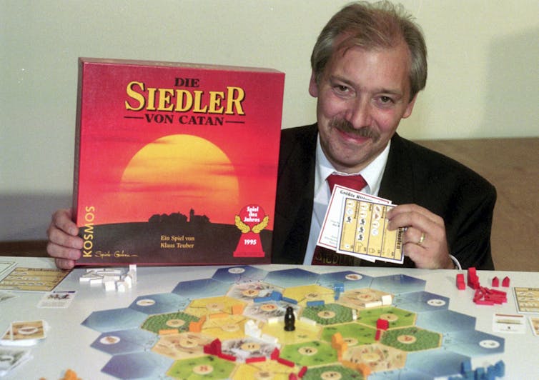 A man wearing a suit holds the Settlers of Catan board game. The game map is on a table in front of him.