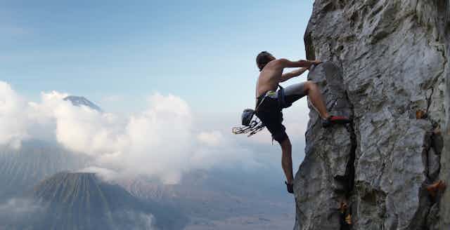 With mountains, clouds and a blue sky in the background, a young man climbs a natural rock wall.