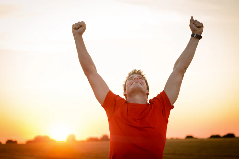 A young man raises his fists in the air and smiles, he is outside at sunset