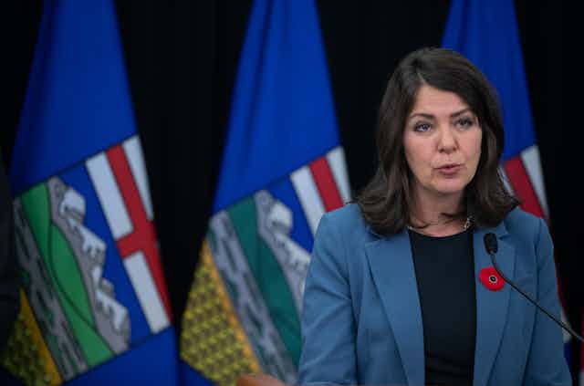 A woman with dark hair sits in front of a row of Alberta flags.