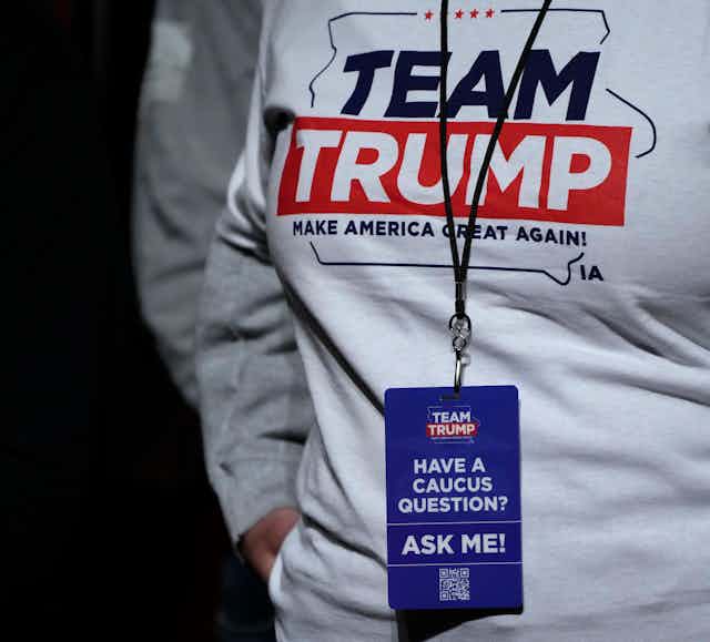 A person wearing a t-shirt that says 'Team Trump,' also wearing a lanyard to which is affixed a card saying 'Have a caucus question? Ask me!'