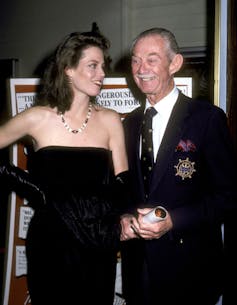 Elderly man with mustache wearing suit smiles as he stands next to a young woman in black dress, who gazes his way.