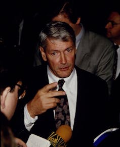 Man with graying hair wearing suit being interviewed.