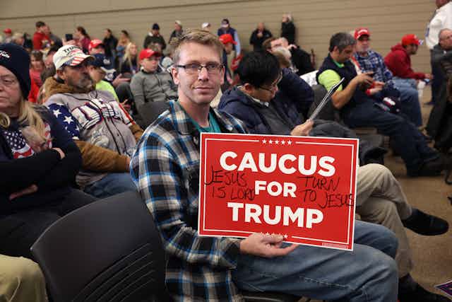A white man is sitting with other people and holds a poster urging people to caucus for Trump.