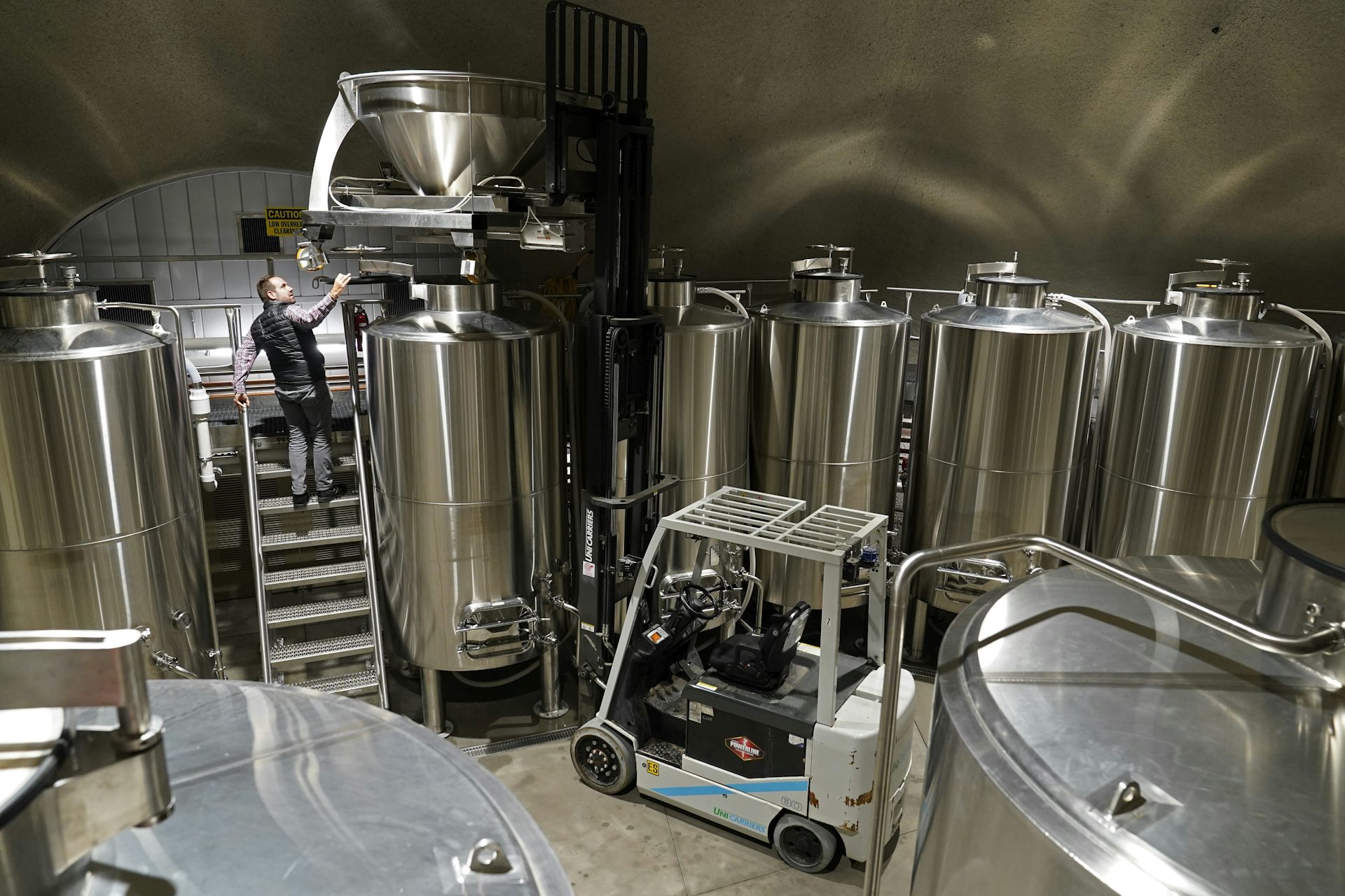 A man stands on a ladder amongst liquid storage tanks and a forklift.