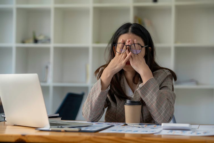 A stressed woman with her face in her hands sitting at a desk with a laptop and papers.