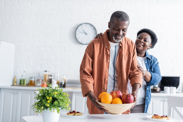 A couple in a kitchen setting a bowl of fruit on the table
